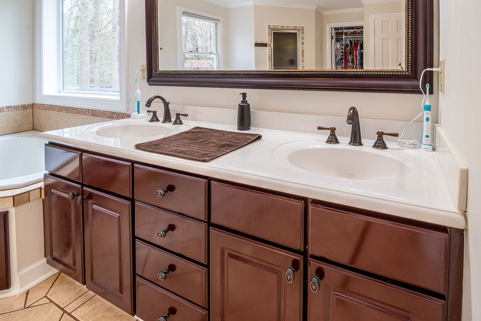 Double vanity with dark wooden cabinets and a large mirror, on the counter a brown towel, a soap dispenser and two electric toothbrushes with a charging cable.