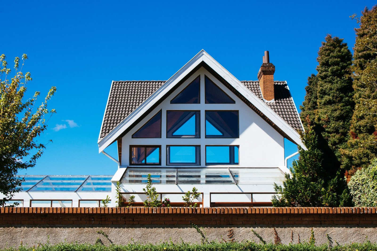 White house with large gable glazing, a dark tiled roof and a chimney behind a garden wall, in daylight under a deep blue sky.