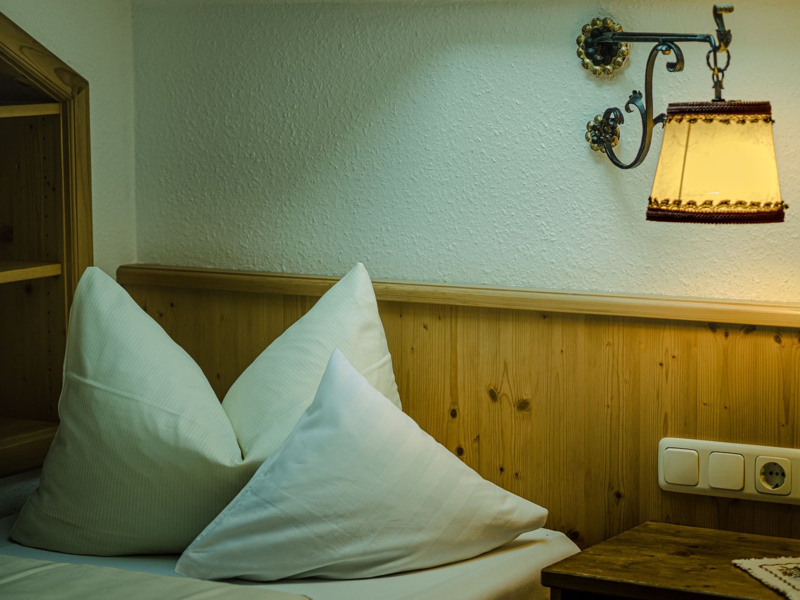 Bed with white pillows against pine panelling, a wrought-iron wall lamp above, wall and pillows with a green cast.