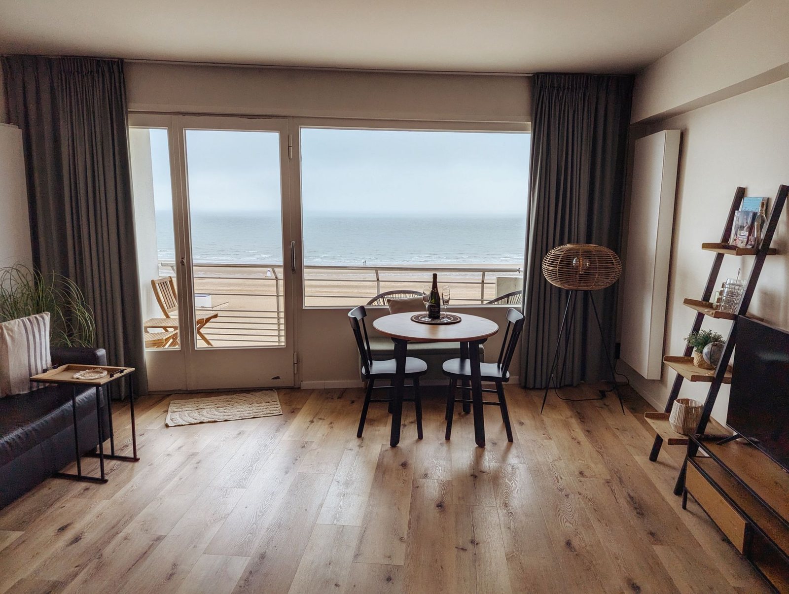 Living room of a holiday flat looking through a glass front and balcony door onto beach and sea, a round table with two chairs and a wine bottle, floor lamp and ladder shelf, flat, cool light.