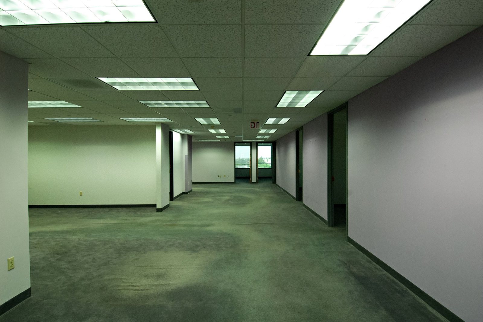 Empty office floor with a grid ceiling, fluorescent panels and carpet, with a green cast and dark towards the back.