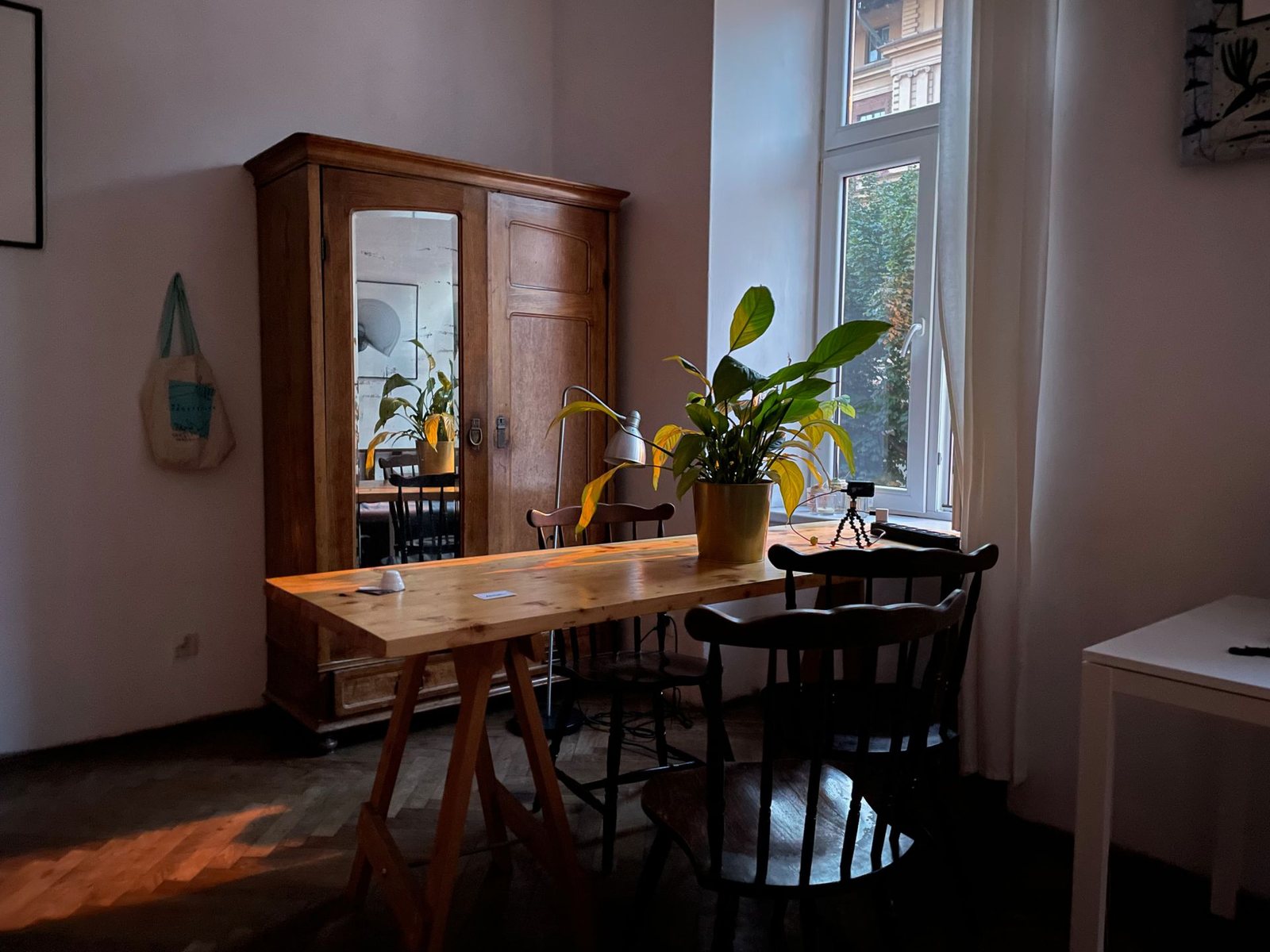 Altbauzimmer mit hohem Fenster, Holzschrank mit Spiegeltür, Holztisch mit Grünpflanze und dunklen Stühlen, zu dunkel, mit Farbstich und leicht gekippten Kanten.