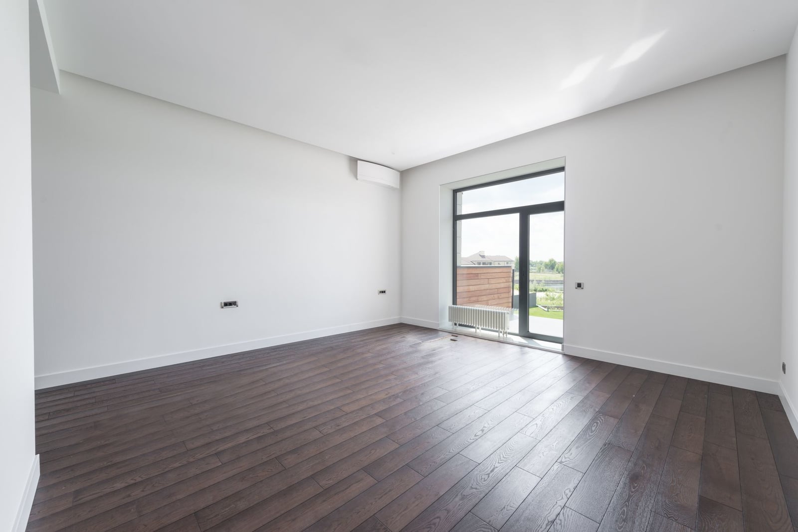Empty living room with dark parquet and a balcony door, no furniture.