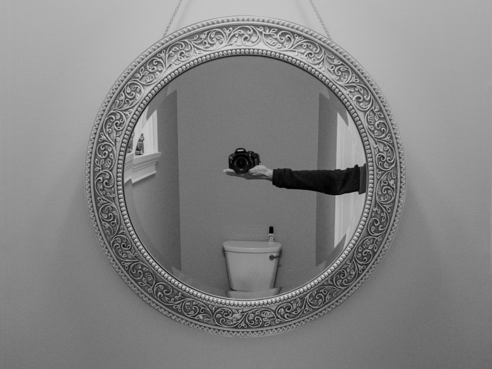 Black-and-white shot of a round wall mirror with an ornate frame, reflected in it an outstretched arm holding a camera, behind it a window sill and a toilet cistern.