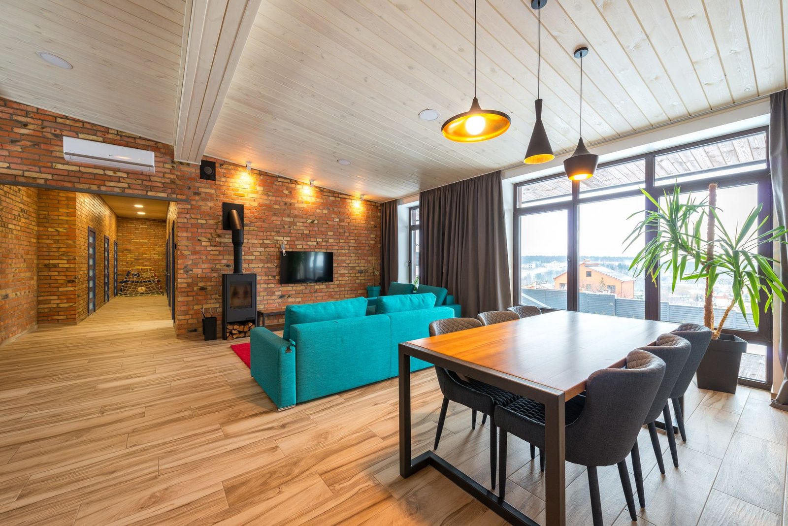 Offener Wohn- und Essbereich mit Ziegelwand, Holzdecke, türkisem Sofa, Kaminofen und großer Fensterfront, im Weitwinkel gekippt aufgenommen, Wände und Fensterrahmen stehen schräg.